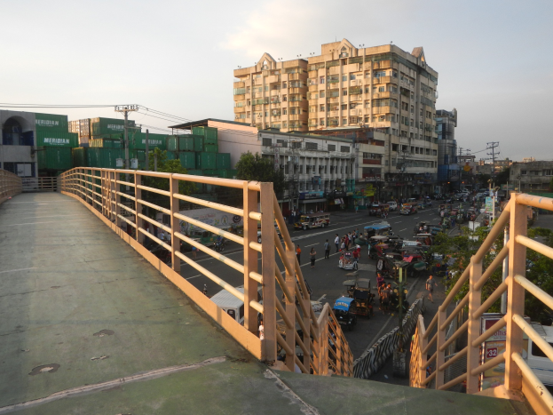 Eine Fußgängerbrücke mit Geländern, die eine stark befahrene Straße überspannt, mit Gebäuden, Bäumen, Strommasten mit Kabeln und einem bewölkten Himmel im Hintergrund.