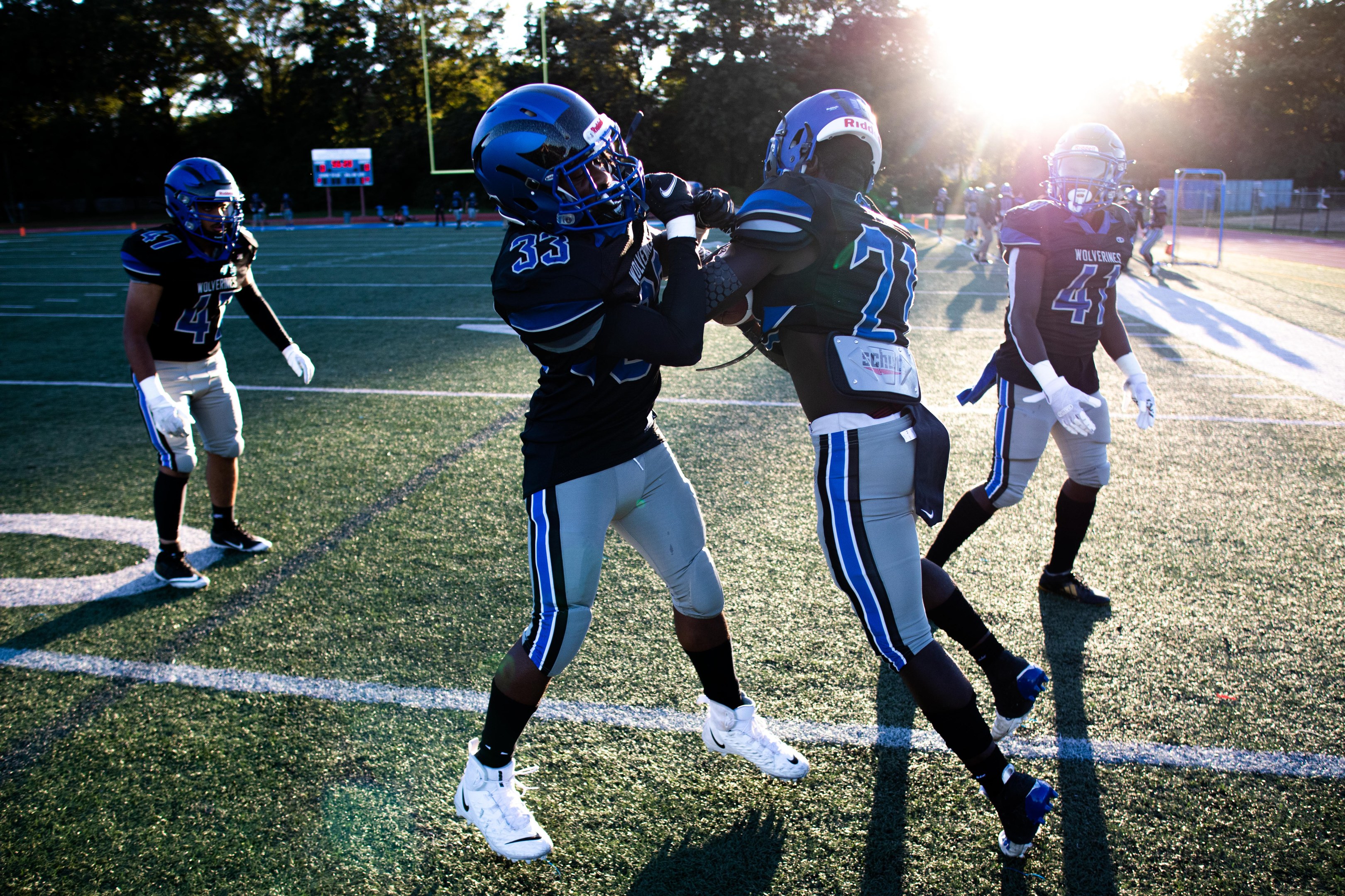 Junge Männer in Helmen und Stollenschuhen spielen Fußball auf einem Feld mit Bäumen und einem klaren blauen Himmel.