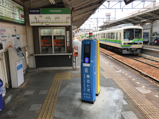 Ein Zug fährt in einen Bahnhof neben einer Plattform mit einigen sitzenden Menschen, Metallstangen, einem deckenmontierten Bildschirm und einem Automaten vor dem Zug.