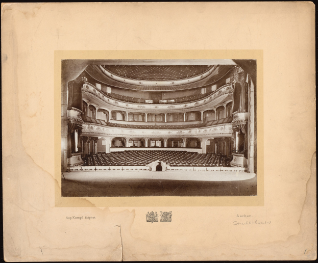 Altes Schwarz-Weiß-Foto des Auditoriums der Royal Albert Hall, das die Bühne, Reihen von Stühlen, Säulen, Geländer und Text mit einem Logo unten zeigt.