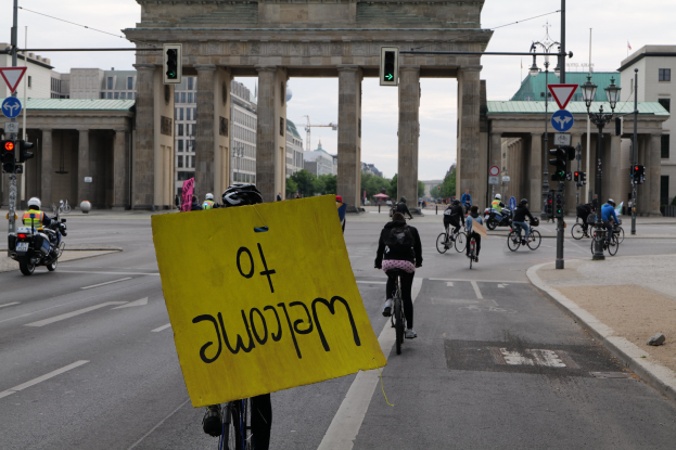 Eine Gruppe von Radfahrern fährt am Brandenburger Tor in Berlin vorbei, einer hält ein gelbes Schild, mit Laternenmasten, Verkehrsampeln, Gebäuden, Bäumen und einem klaren Himmel im Hintergrund.