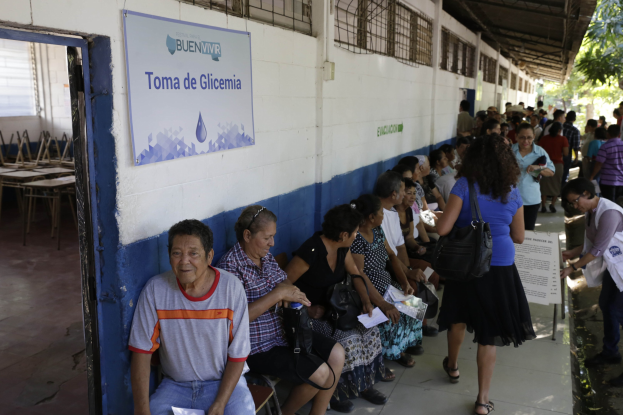 Eine Gruppe von Menschen sitzt auf Bänken vor einem Gebäude, einige halten Papiere und Taschen, mit einem Textschild an der Wand dahinter, Tischen und Stühlen links und Bäumen und einem Schuppen im Hintergrund.