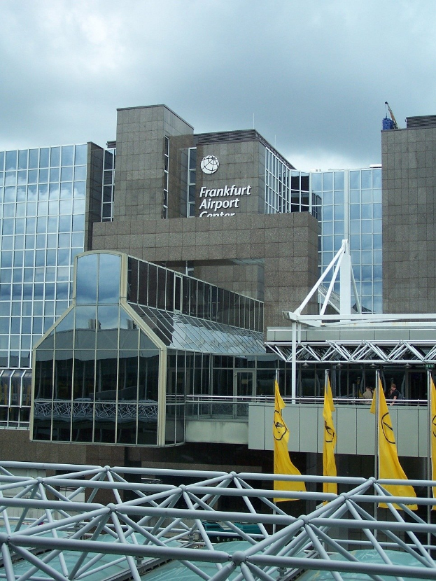 Frankfurt Airport Gebäude mit Glaswänden, Text, gelben Fahnen und Eisenstangen unter einem bewölkten Himmel.