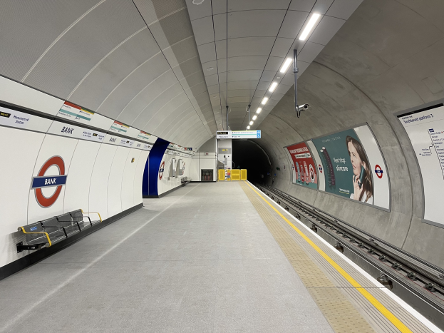 Eine U-Bahn-Station mit einem Zug auf den Gleisen, eine Plattform mit Bänken, Tafeln mit Text und Bildern, Lampen an der Decke und eine Überwachungskamera.