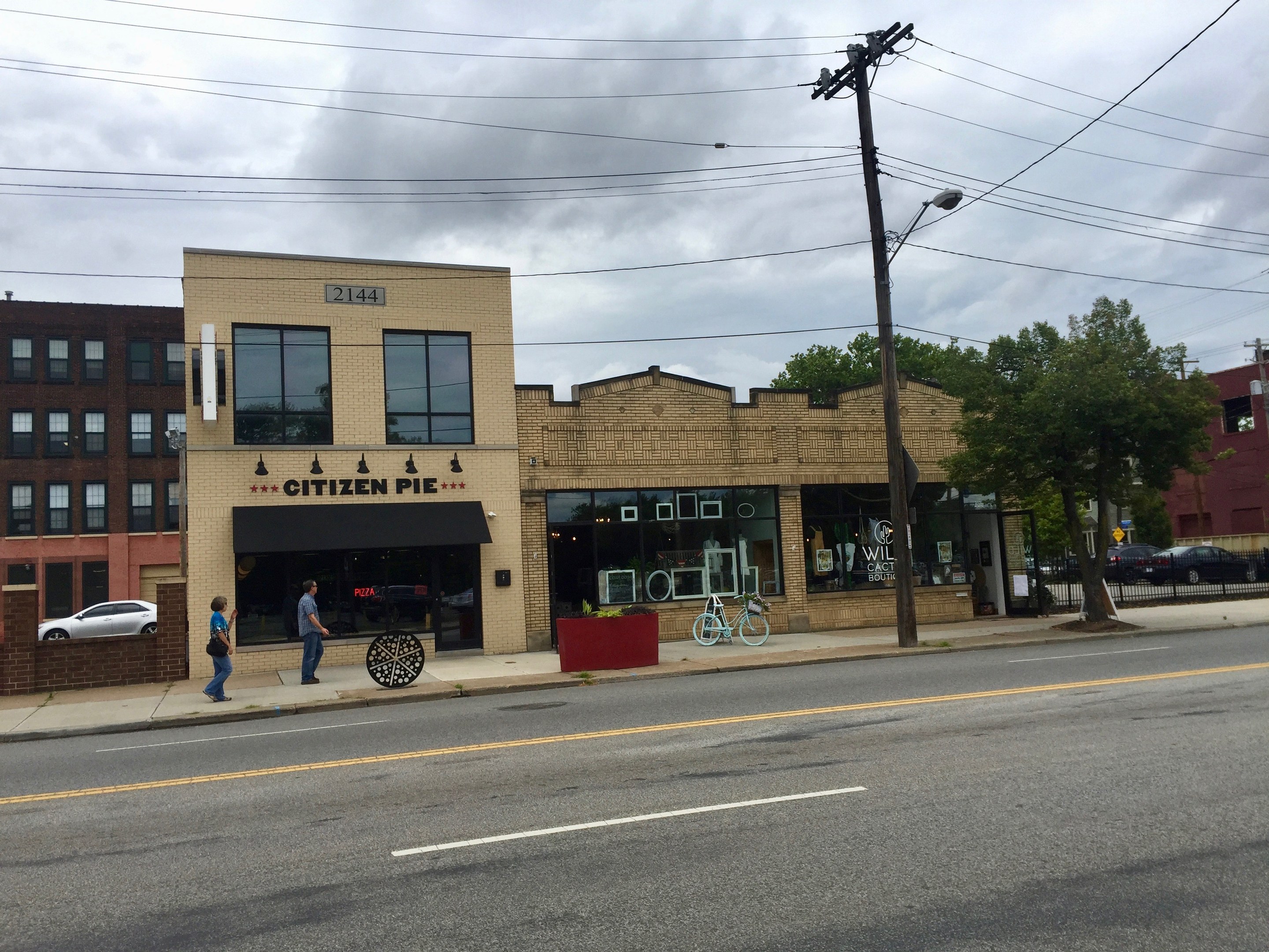 Eine belebte Stadtstraße mit Fußgängern, parkenden Fahrrädern, Strommasten, Bäumen und Gebäuden, mit dem Restaurant Citizen Pie an der Ecke unter einem bewölkten Himmel.