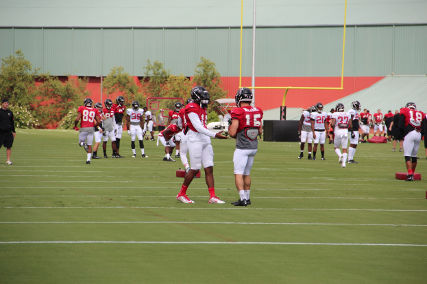 Gruppe von Menschen, die Fußball auf einem Rasenfeld mit Helmen und Stiefeln spielen, umgeben von Bäumen, Pfählen, einer Wand und einem klaren blauen Himmel.