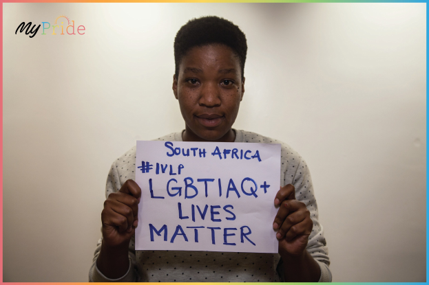 Eine Frau in einem weißen Hemd steht entschlossen vor einer Wand und hält ein Schild mit der Aufschrift "LGBTQ+ Lives Matter".