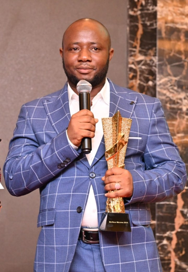 Ein Mann in einem blauen Anzug hält ein Mikrofon und eine Trophäe und steht selbstbewusst mit einem freudigen Gesichtsausdruck vor einer Wand.