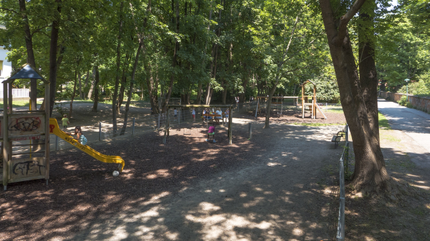 Ein Spielplatz in einem Park, umgeben von Bäumen, mit Menschen auf den Geräten und einem Gebäude links daneben.