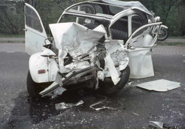 Ein schwerbeschädigt auf der Fahrbahn stehendes Auto mit Frontalaufprall, umgeben von Bäumen und einem entfernten Gebäude, mit einem weiteren Fahrzeug im Hintergrund.