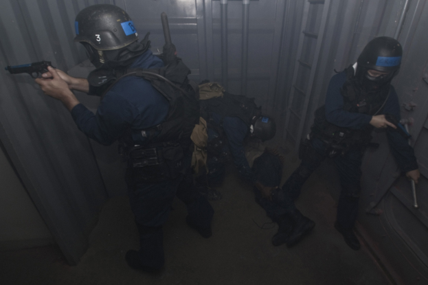 Eine Gruppe von Polizisten in Einsatzausrüstung, einer mit einer Waffe in der Hand, neben einem Metallcontainer und einer Leiter in einem begrenzten Raum.