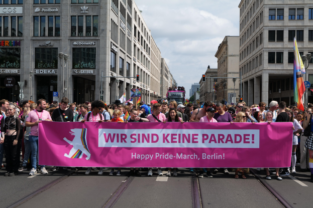 Eine Gruppe von Menschen marschiert auf einer Straße in Berlin, Deutschland, mit einer pinken Fahne, auf der 'Happy Pride March' steht, vorbei an Gebäuden, Laternenmästen und Ampeln.