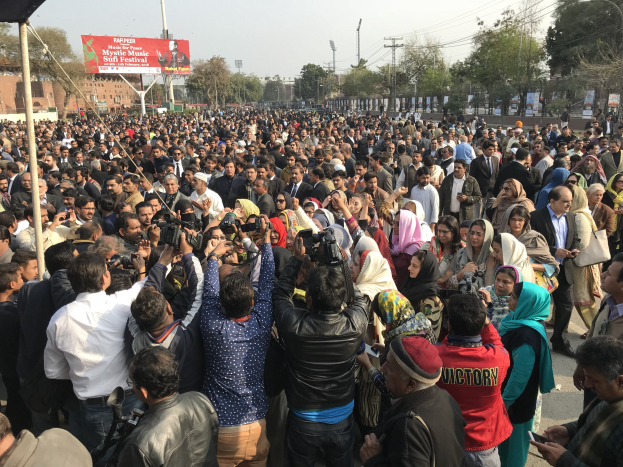 Eine große Menschenmenge versammelt sich vor einem Gebäude, einige halten Kameras und Telefone, mit einem Banner mit der Aufschrift "Kashmiri-Muslime protestieren in Lahore" zwischen Metallrahmen, Pfählen und Bäumen unter einem bewölkten Himmel.
