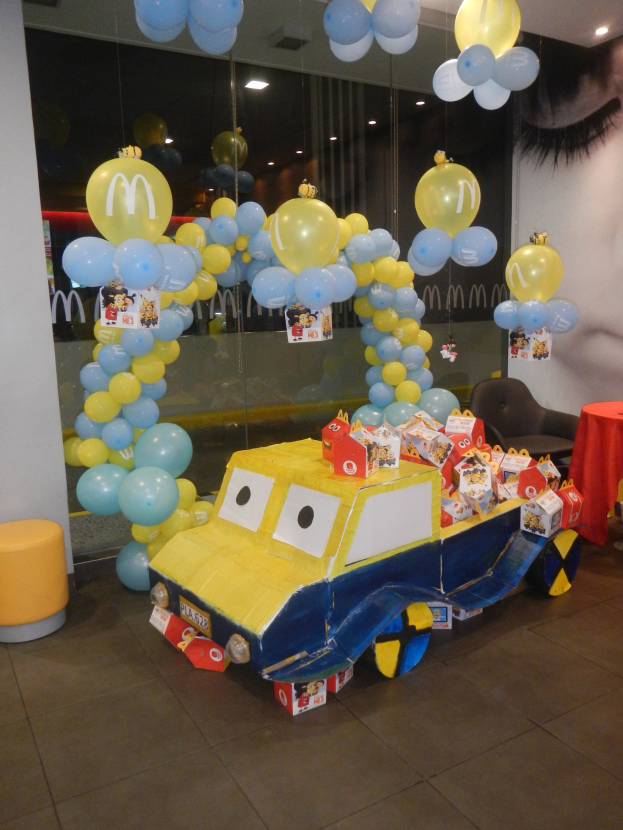 Eine McDonald's-Geburtstagsfeier mit einem gelben und blauen Lastwagen, der mit Ballons und anderen Dekorationen geschmückt ist, einem rot gedeckten Tisch mit Gegenständen, Hockern und einer Glaswand im Hintergrund unter festlicher Deckenbeleuchtung.