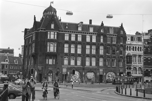 Ein Schwarz-Weiß-Foto von Menschen, die auf der Straße Fahrräder fahren, umgeben von Gebäuden, Straßenlaternen, Verkehrspfosten, Verkehrsampeln, Kraftfahrzeugen, Fußgängern auf dem Gehweg, Absperrpollern, Stromkabeln und Himmel.