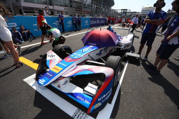 Formula E-Rennwagen mit weißer, blauer, roter und schwarzer Lackierung auf einer Rennstrecke geparkt, umgeben von Menschen, mit einem Schirm im Vordergrund und Infrastruktur am Rand der Strecke im Hintergrund bei klarem Himmel.
