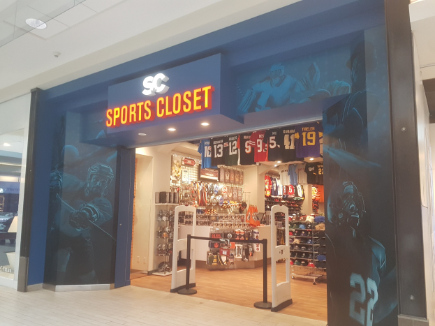 Ein Sportbekändler im Einkaufszentrum mit einem "SC Sports Closet"-Schild, das Sportbekleidung auf Bügeln und organisierte Gegenstände in Schubladen unter Deckenlampen zeigt, mit einer gläsernen Eingangstür auf der linken Seite.
