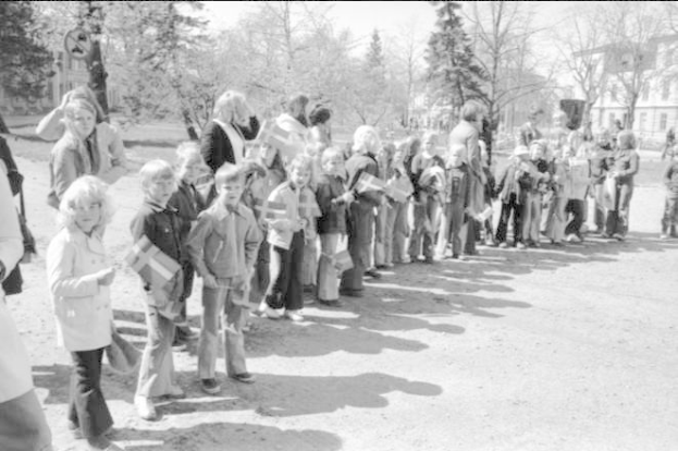 Ein Schwarz-Weiß-Bild einer Gruppe von Menschen, die in einer Reihe auf einem Schotterweg stehen, Fahnen halten, mit Bäumen, Gebäuden und einem klaren Himmel im Hintergrund, die an einer Protestaktion auf dem Schulgelände teilnehmen.