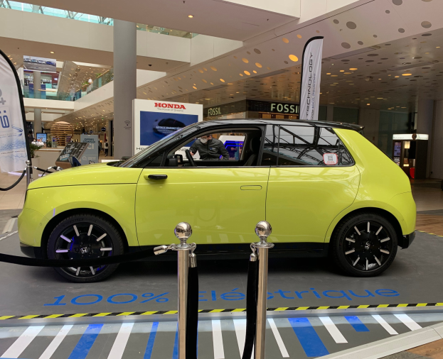 Gelbes wasserstoffbetriebenes Elektroauto vor einem Gebäude mit Absperrpollern, Bannern, Menschen, Schildern, einer Topfpflanze, Deckenleuchten und Ventilatoren.