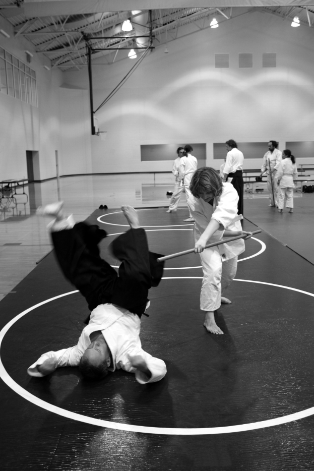 Zwei Personen in einer Turnhalle üben Karate mit Stöcken, während andere zusehen, mit Wandbeleuchtung im Hintergrund.
