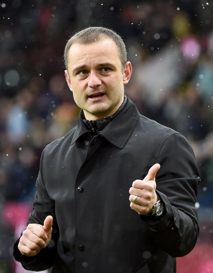 Ein Mann in einer schwarzen Jacke zeigt den Daumen hoch, trägt eine Uhr und hat einen unscharfen Hintergrund.