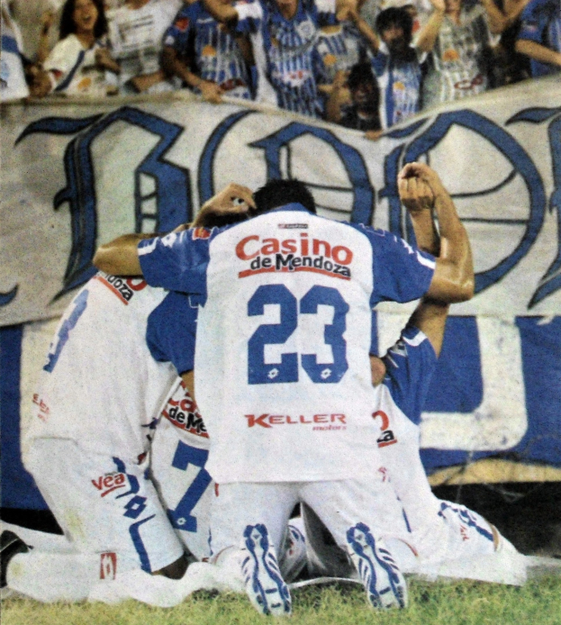 Fußballspieler feiern ein Tor auf dem Feld mit jubelnden Zuschauern im Hintergrund und einem Banner mit Text dahinter.