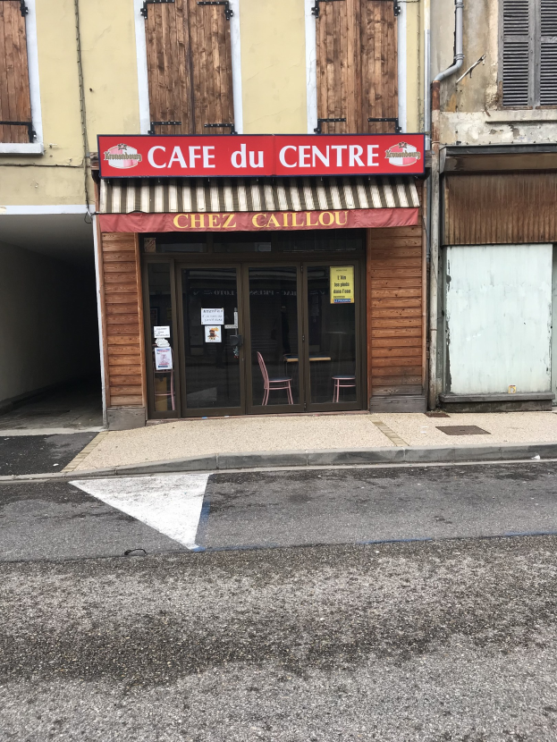 Außenansicht eines Gebäudes mit einer 'Cafe du Centre'-Tafel, einer Straße davor, einer Tafel mit Text, Fenstern und einer Tür.