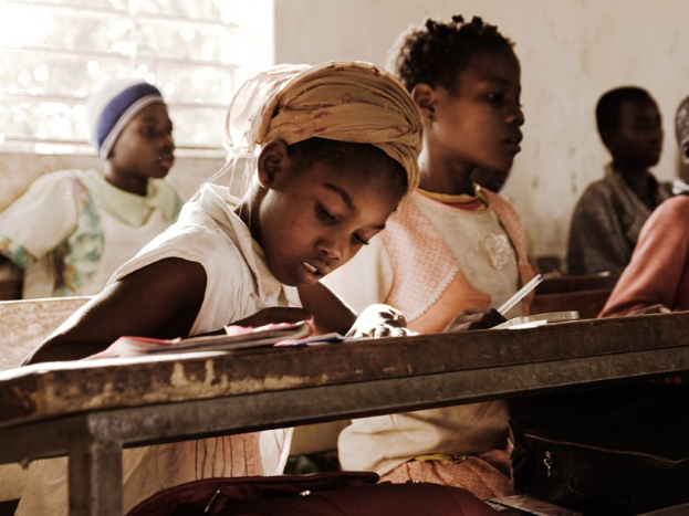 Eine Gruppe von Kindern sitzt an Tischen in einem Klassenzimmer, mit Büchern und Taschen vor ihnen auf den Bänken und einem Fenster im Hintergrund.