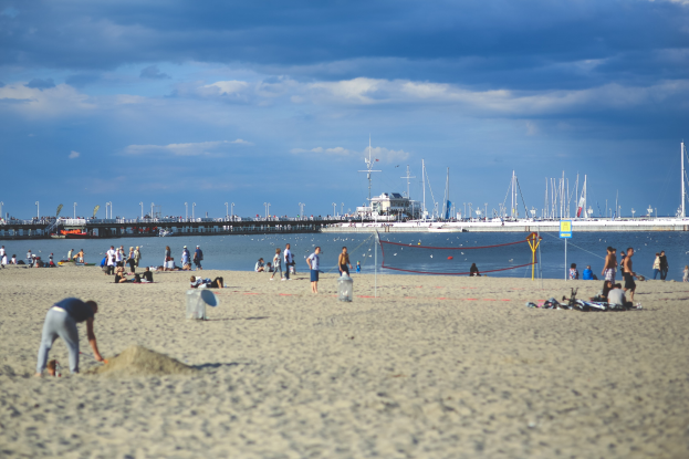 Eine Gruppe von Menschen, die Volleyball am Strand spielen, mit einem Netz, das auf einen See mit Booten, Pfählen, eine Brücke und einen bewölkten Himmel blickt.