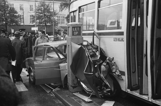 Schwarz-weißes Bild eines Bus, der auf der Seite der Straße in ein Auto gekracht ist, mit Zuschauern und Gebäuden im Hintergrund.