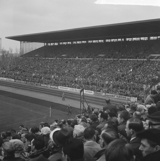 Schwarzes und weißes Foto eines vollen Stadions mit Zuschauern, die ein Fußballspiel beobachten, einige stehen und andere sitzen auf den Tribünen, mit Bannern, Pfählen, einem Schuppen, Bäumen, einem Turm und einem bewölkten Himmel im Hintergrund.