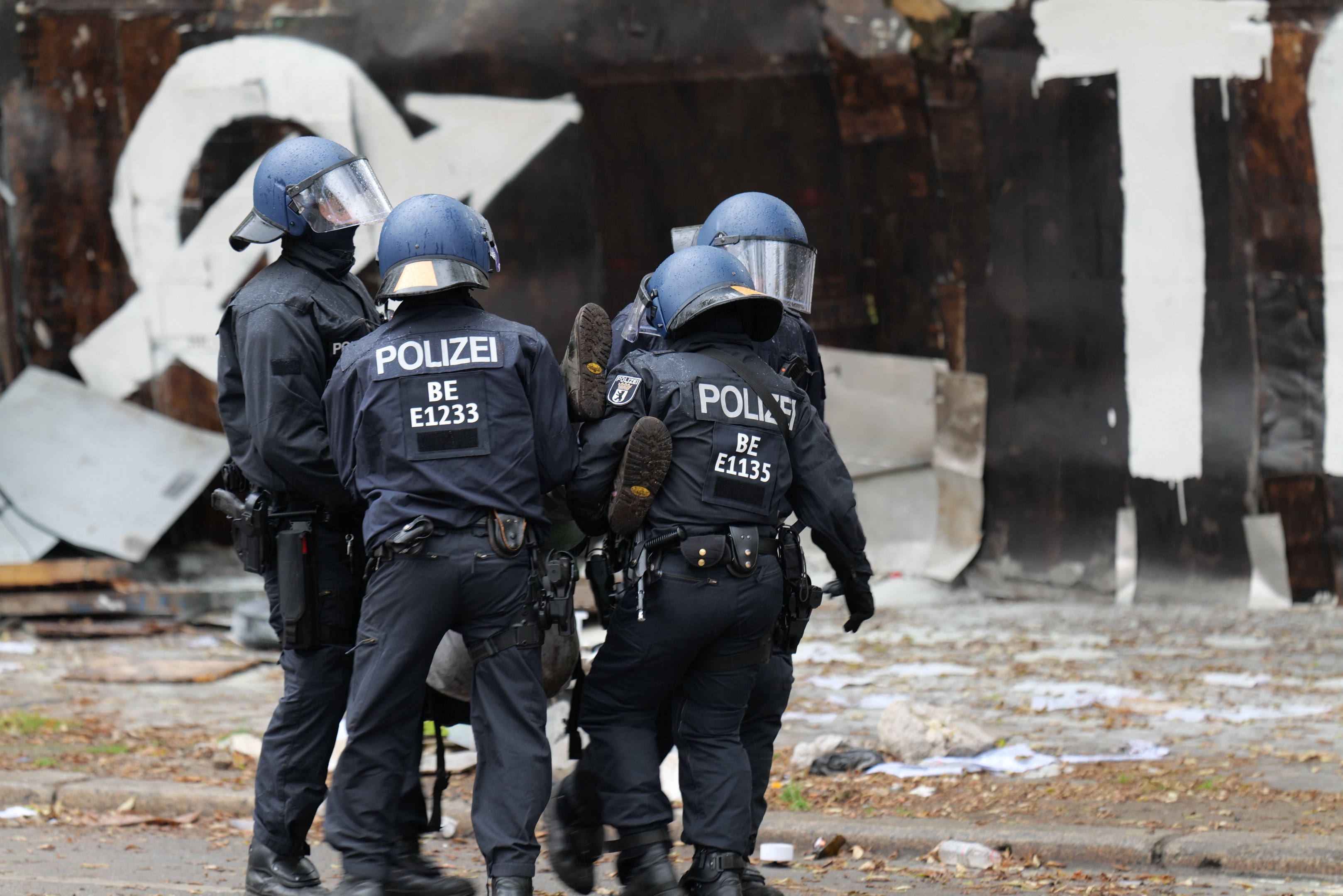 Eine Gruppe von Polizisten in schwarzen Uniformen und blauen Helmen steht vor einem Gebäude mit Graffiti, umgeben von verstreuten Papieren und Gegenständen.