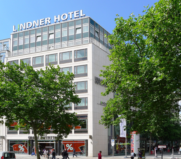 Großes gläsernes Gebäude mit der Aufschrift "Lindner Hotel" in Berlin, umgeben von Bäumen, Fahrzeugen und Passanten unter einem sichtbaren Himmel.