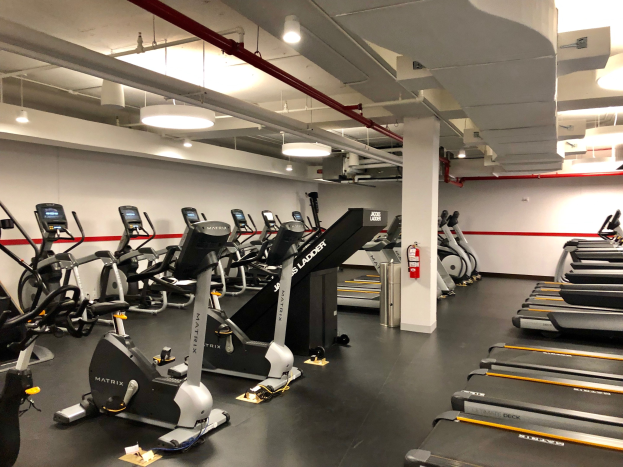 Ein Fitnessstudio in New York City mit einer Reihe von Laufbändern und Ellipsentrainern, einer Säule, einem Feuerlöscher an der Wand und Deckenleuchten.