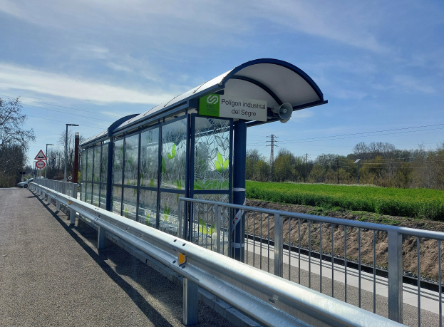 Ein Bushaltestellenhäuschen mit einem Dach, Schildern und Straßenmöbeln an einer Straße, umgeben von Bäumen und Pflanzen unter einem bewölkten Himmel.