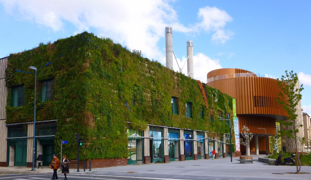 Ein großes Gebäude mit einer grünen Wand an der Seite, umgeben von Straßenlaternen, Verkehrspfosten, Verkehrssignalen, Kraftfahrzeugen, Fußgängern, Bäumen und einem bewölkten Himmel als Teil des Green Building Projects.
