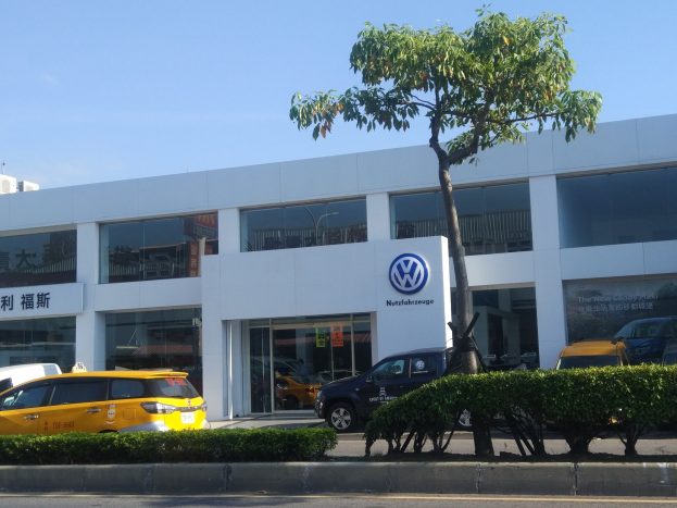 Ein VW-Autohaus mit parkenden Autos vorne, umgeben von Pflanzen und einem Baum, mit Glaswänden und Fenstern unter einem sichtbaren Himmel.
