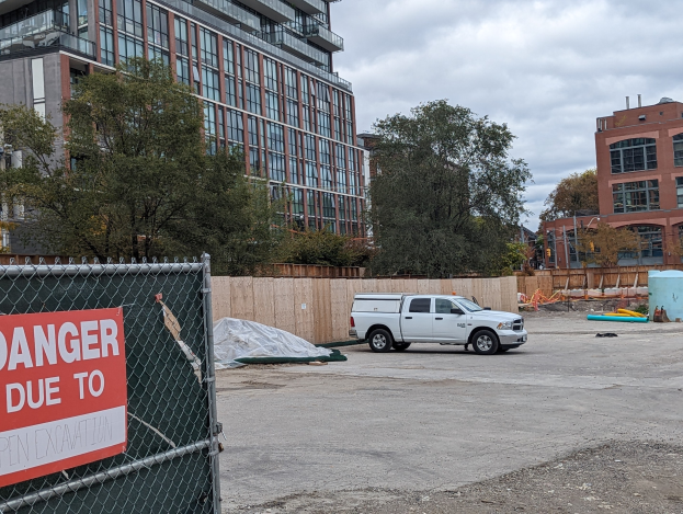 Lastwagen vor einem Gebäude mit einer "Gefahr wegen offener Baustelle"-Tafel geparkt, mit Bäumen, Gebäuden, Polen und einem klaren blauen Himmel im Hintergrund.