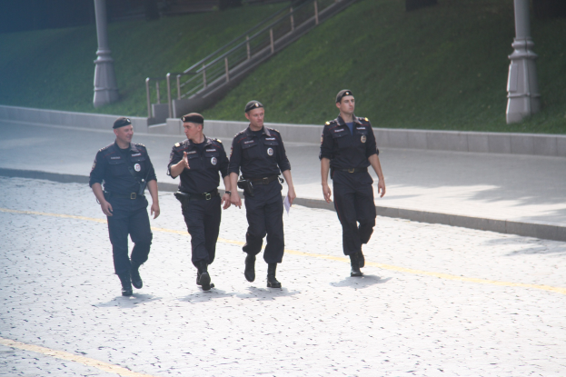 Drei Polizisten in Uniform und mit Mützen, die eine Straße mit Pfählen, Geländern, Treppen und Gras im Hintergrund entlanggehen.