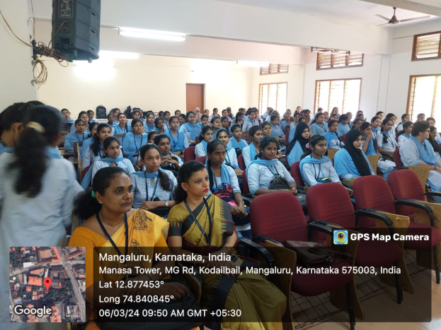Eine Gruppe von Menschen sitzt in Stühlen in einem Raum mit einer Wand, Fenstern auf der rechten Seite, Deckenlampen und einem Ventilator und einem Redner auf der linken Seite, mit Text und einer Karte unten.