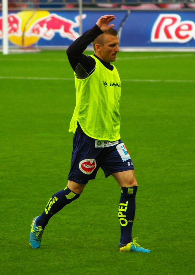 Ein Mann in einem gelben T-Shirt und blauen Shorts steht auf einem Fußballfeld bereit zum Spiel, umgeben von einem Netz, Werbetafeln und Stühlen.