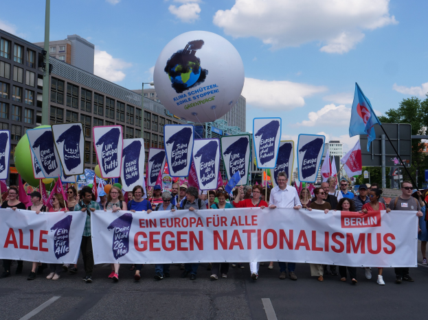 Eine Gruppe von Menschen marschiert die Straße entlang, hält ein Banner mit der Aufschrift "Ein Europa für alle gegen Nationalismus" und Fahnen, mit Gebäuden, Bäumen und einem bewölkten Himmel im Hintergrund.