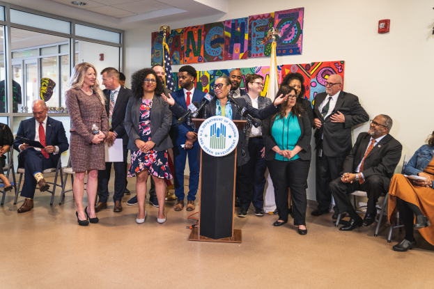 Eine Gruppe von Menschen, die vor einem Podium mit Mikrofon und einer Tafel mit Text darauf stehen und sitzen, mit Flaggen, einer Tafel, einem Feuerlöscher und Deckenlampen an der Rückwand.
