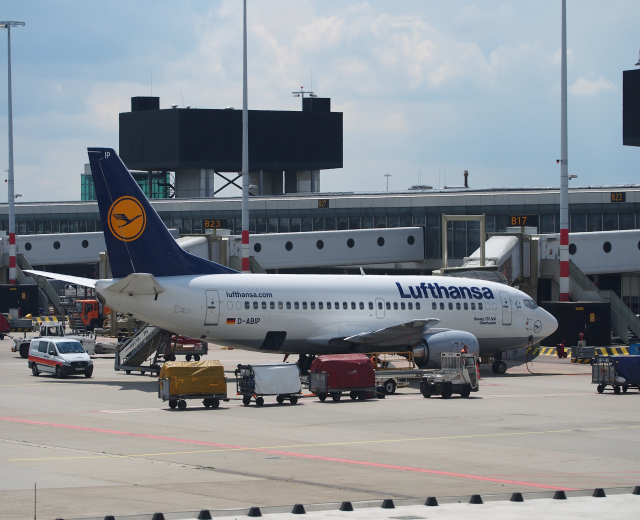 Lufthansa Airbus A320-200 am Frankfurter Flughafen mit Bodenfahrzeugen und Wagen im Vordergrund, einem Gebäude und Laternen im Hintergrund, unter einem klaren blauen Himmel.