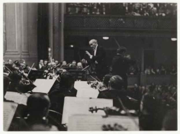 Schwarzes und weißes Foto eines Dirigenten vor einem Orchester, der einen Taktstock hält, mit sitzenden Musikern, die Instrumente spielen und Notenständer mit Büchern im Vordergrund, eine Wand und ein Geländer im Hintergrund.