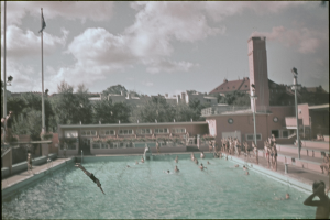Altes Schwarz-Weiß-Foto von Menschen, die in einem Pool schwimmen, mit Gebäuden, Bäumen, Polen, Flaggen und einem Uhrenturm im Hintergrund unter einem bewölkten Himmel.