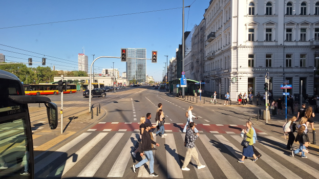 Gruppe von Menschen, die eine Straße an einer Ampel überqueren, mit einem Bus auf der linken Seite, Fahrzeugen auf der Straße, Verkehrsampeln, Laternen, Gebäuden, Bäumen und einem klaren blauen Himmel.