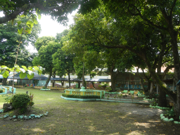 Ein Park mit grünem Gras, Pflanzen, Bäumen und einem zentralen Brunnen, vor Buildings und einem klaren blauen Himmel.