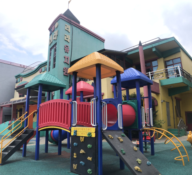 Buntes Kinderspielgerät vor einem Gebäude mit Fenstern, umgeben von Bäumen und einem bewölkten Himmel.