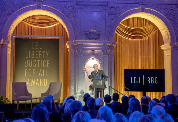 Ein Mann steht an einem Rednerpult mit einem Mikrofon, das eine sitzende Menge in einem Raum mit Stühlen, Pflanzen, Vorhängen und einer Tafel mit Text dahinter anspricht.